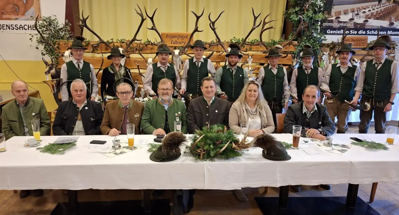 Eine Gruppe von Menschen in traditioneller Kleidung posiert für ein Foto an einem Tisch mit Biergläsern und Geweihen. Sie stehen vor einer Holzwand mit einem Schild, das 'Eigenjagd Isbary' steht.