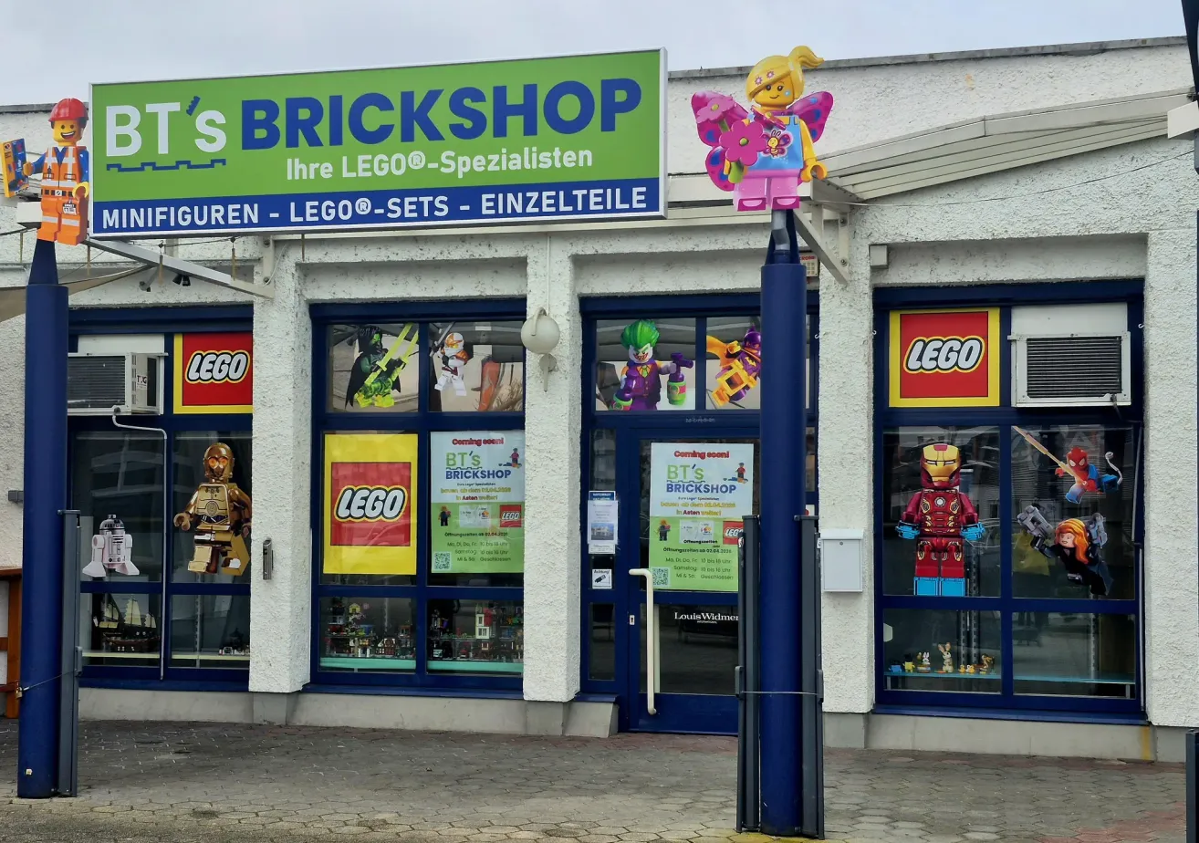 Außenansicht eines LEGO-Spezialistengeschäfts. Das grüne Schild trägt die Aufschrift 'S BRICKHSHOP'. Im Schaufenster sind LEGO-Charaktere wie Iron Man und eine LEGO-Figur mit Blumen zu sehen.