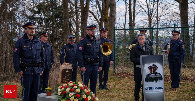 Eine Gedenkfeier mit Polizeibeamten, die um ein Grab stehen, einer von ihnen spricht an einem Podium, mit einem Banner hinter ihm.