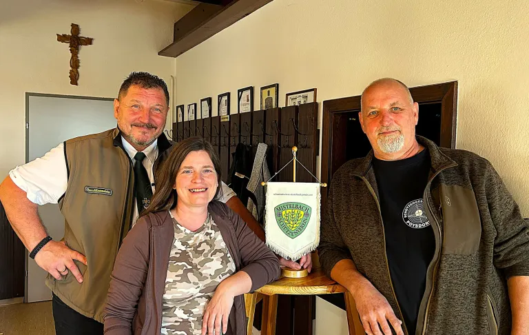 Drei Personen stehen in einem Raum mit einer Flagge und einem Banner. Eine Frau in der Mitte trägt ein Camouflage-Shirt. Der Mann auf ihrer linken Seite trägt einen braunen Mantel und eine Krawatte. Der Mann auf ihrer rechten Seite trägt einen braunen Mantel.