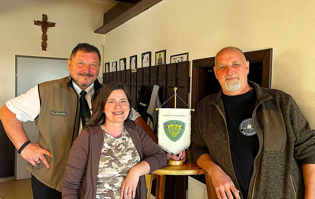 Drei Personen stehen in einem Raum mit einer Flagge und einem Banner. Eine Frau in der Mitte trägt ein Camouflage-Shirt. Der Mann auf ihrer linken Seite trägt einen braunen Mantel und eine Krawatte. Der Mann auf ihrer rechten Seite trägt einen braunen Mantel.
