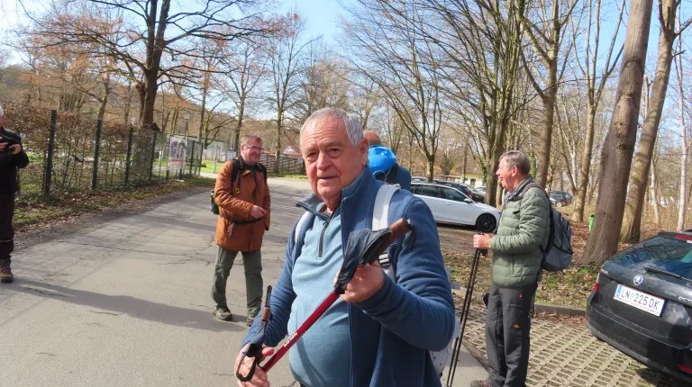 Eine Gruppe von Männern, wahrscheinlich Wanderer, geht auf einem gepflasterten Weg. Der Mann vorne hält eine Trekking-Stange und lächelt. Die Männer hinter ihm halten ebenfalls Trekking-Stöcke. Sie tragen Jacken, Hosen und Turnschuhe. In der Ferne sind Bäume und Autos zu sehen.