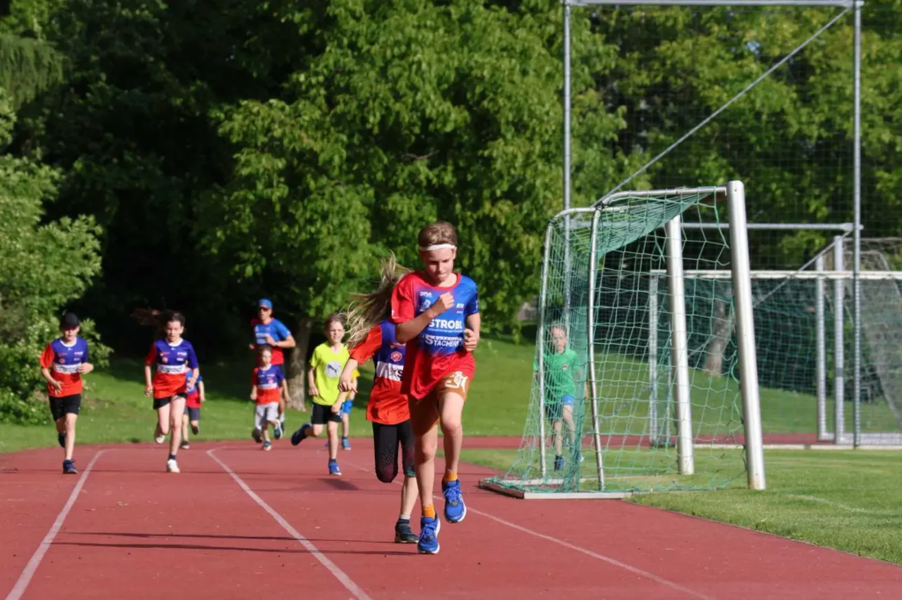 Kinder laufen auf einer Laufbahn, eines trägt ein blaues Hemd mit 'STROB', ein anderes ein rotes Hemd. Ein Fußballtor ist in der Nähe.