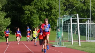 Kinder laufen auf einer Laufbahn, eines trägt ein blaues Hemd mit 'STROB', ein anderes ein rotes Hemd. Ein Fußballtor ist in der Nähe.
