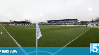 Ein Fußballfeld mit Spielern im Training, blaue Tribünen im Hintergrund und eine weiße Flagge auf einem Pfosten.