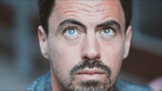 Nahaufnahme eines Mannes mit blauen Augen, Bart und blauem Hemd. Sein Gesicht zeigt Anzeichen des Alterns mit Falten und einem leicht müden Blick.