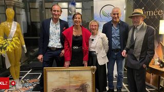 Eine Gruppe von fünf Personen steht gemeinsam in einem Raum, lächelt und posiert für ein Foto mit einem gerahmten Gemälde. Der Mann auf der linken Seite trägt einen Gürtel und einen Anzug, während die anderen lässig gekleidet sind.