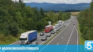 Eine Autobahn in Österreich zeigt einen Stau mit zahlreichen Autos und Lastwagen auf der Straße. Das Gebiet ist von viel Grün und Bergen umgeben.