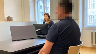 Ein Mann sitzt an einem Schreibtisch vor zwei Monitoren. Eine Frau sitzt hinter ihm und sieht den Mann an. Der Raum hat eine weiße Wand und ein Fenster mit Blick auf ein Gebäude.
