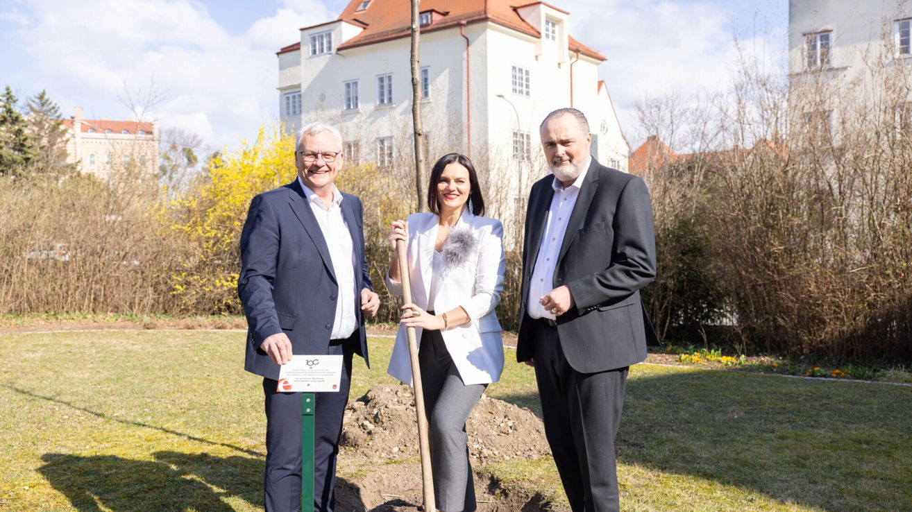 Drei Personen stehen neben einem frisch gepflanzten Baum. Die Frau hält eine Schaufel, und die beiden Männer tragen Anzüge. Sie lächeln und scheinen für ein Foto zu posieren. Dahinter ist ein großes Gebäude zu sehen.