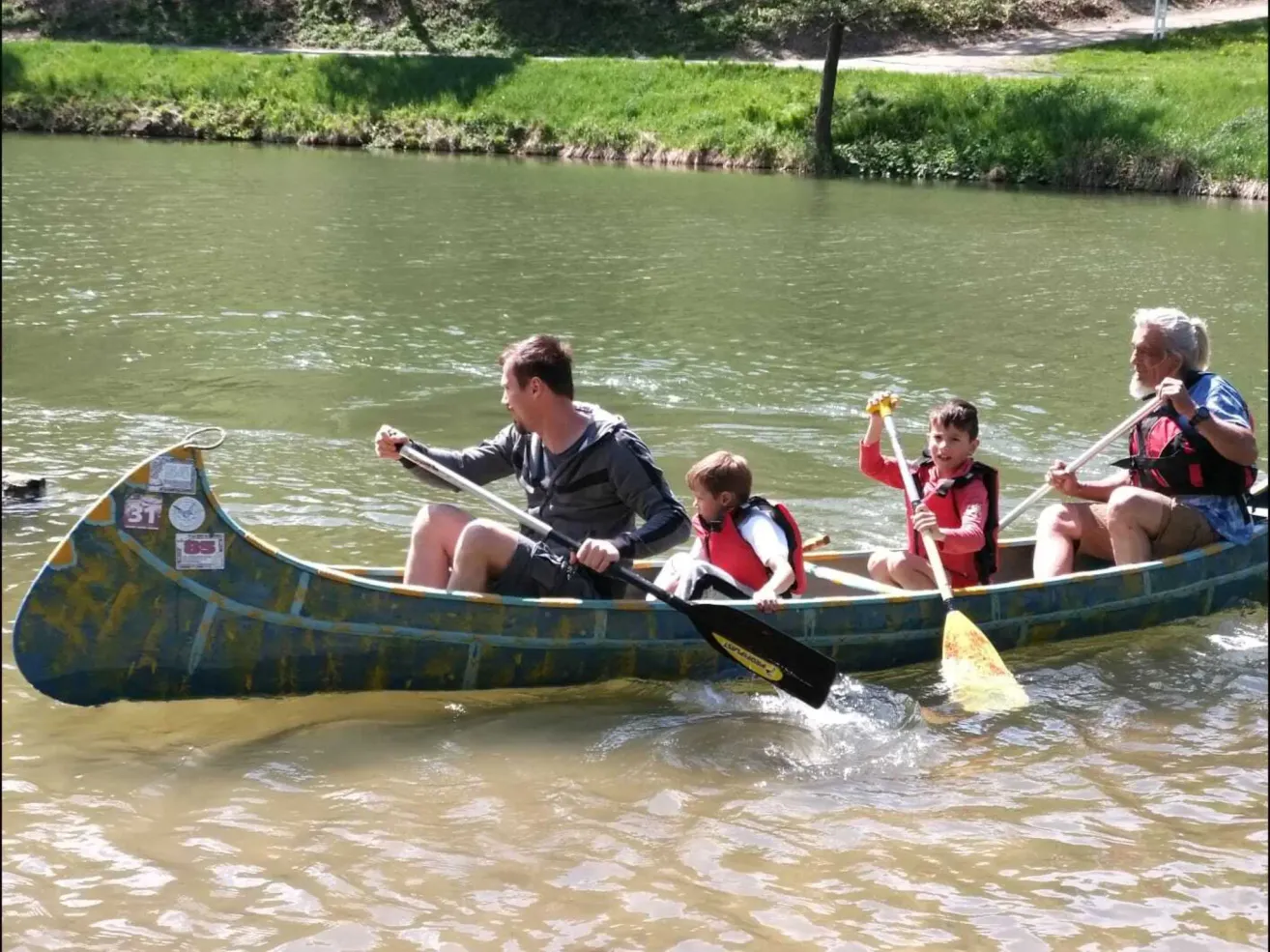 Ein Mann paddelt ein Kanu mit drei Kindern darauf auf einem Fluss mit grasbewachsenen Ufern.
