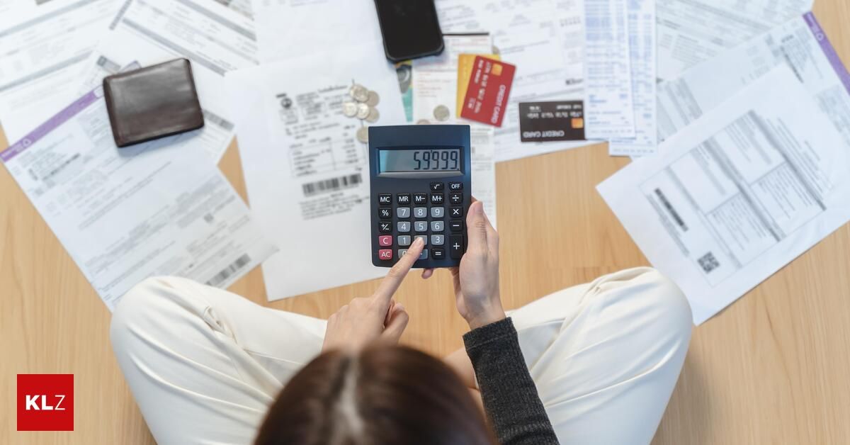 Eine Frau sitzt an einem Schreibtisch und zeigt mit dem Finger auf eine Taschenrechner, der die Zahl 59999 anzeigt. Verschiedene Papiere, Karten, Münzen und ein Handy sind auf dem Schreibtisch verstreut.