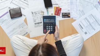 Eine Frau sitzt an einem Schreibtisch und zeigt mit dem Finger auf eine Taschenrechner, der die Zahl 59999 anzeigt. Verschiedene Papiere, Karten, Münzen und ein Handy sind auf dem Schreibtisch verstreut.