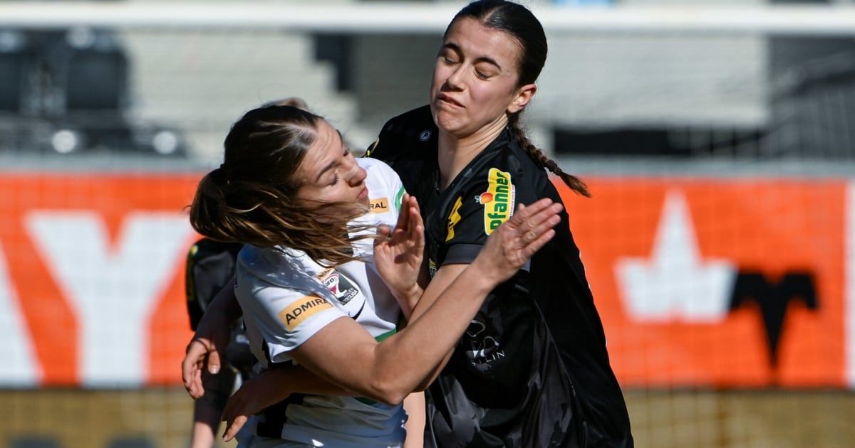Zwei Sportlerinnen in Sportuniformen sind in einem körperlichen Wettkampf auf einem Feld. Die Frau in Weiß hat die Augen geschlossen, während sie von der Frau in Schwarz festgehalten wird.