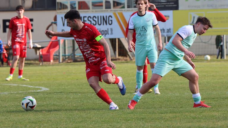 Ein Fußballspieler in roter Uniform läuft mit dem Ball, verfolgt von einem Gegner in hellgrüner Uniform. Dahinter beobachtet ein weiterer Spieler in rot-weißer Uniform.