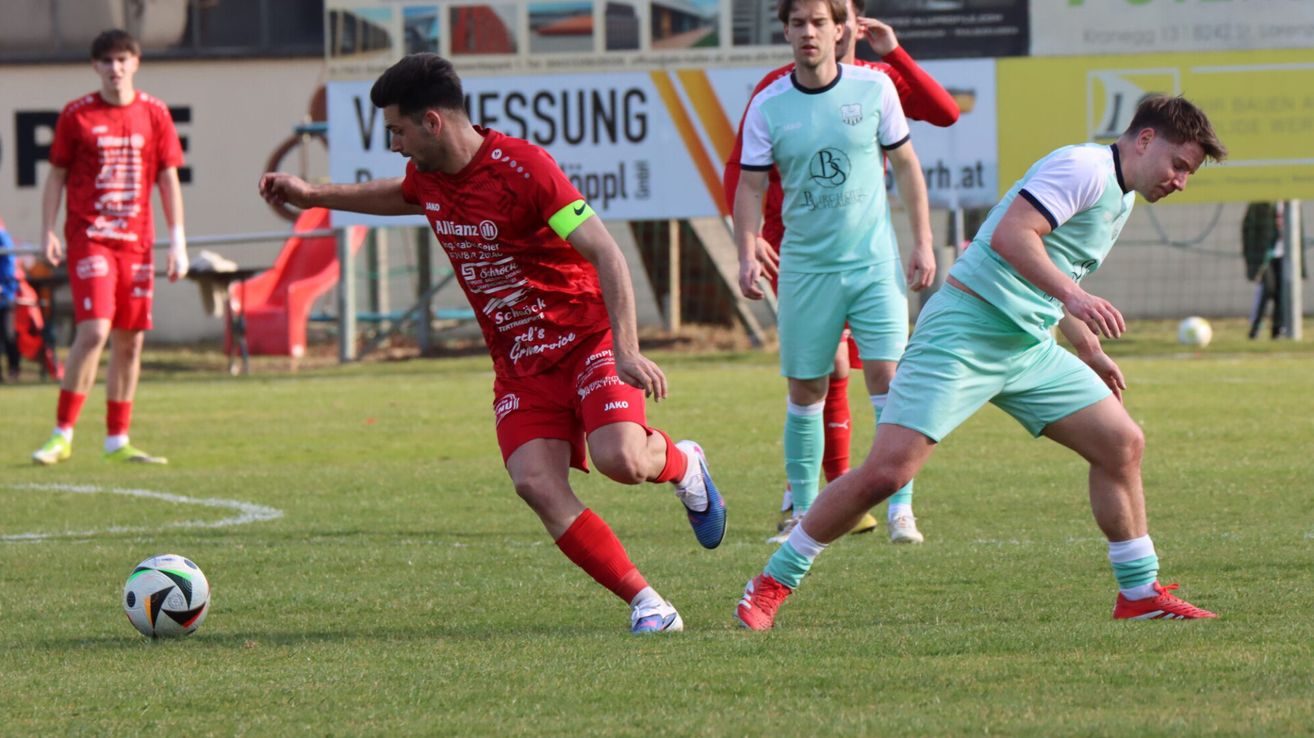 Ein Fußballspieler in roter Uniform läuft mit dem Ball, verfolgt von einem Gegner in hellgrüner Uniform. Dahinter beobachtet ein weiterer Spieler in rot-weißer Uniform.