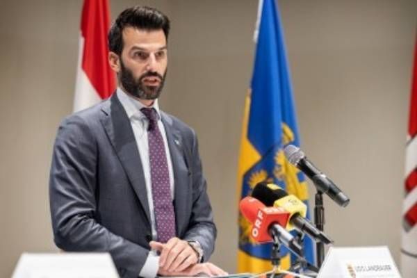 Ein Mann in einem grauen Anzug steht an einem Rednerpult und hält eine Pressekonferenz mit Fahnen hinter ihm ab.