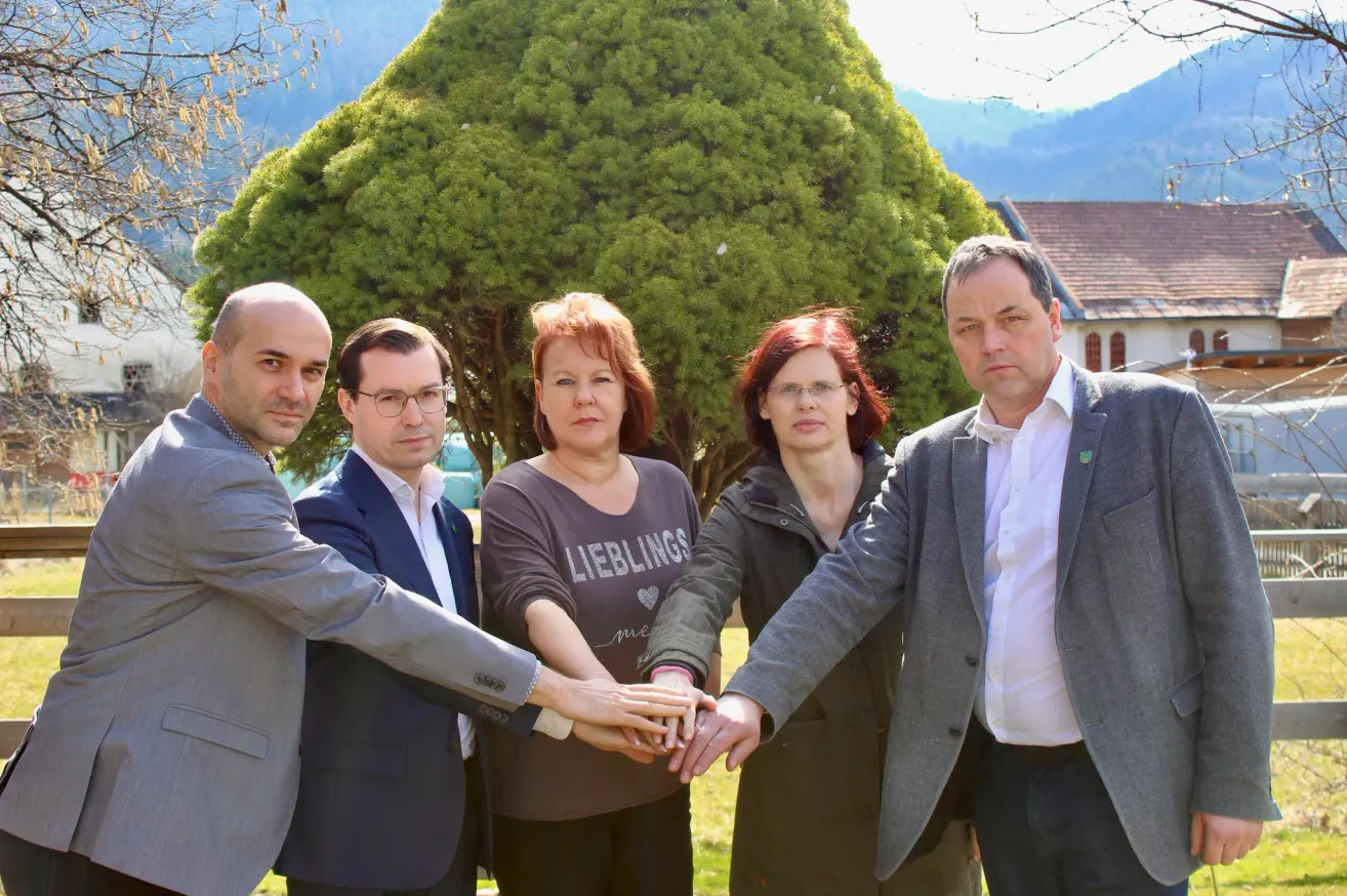 Fünf Personen stehen in einer Reihe, Hände vor einem großen Baum mit Bergkulisse. Zwei Frauen und drei Männer, alle formal gekleidet.