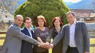 Fünf Personen stehen in einer Reihe, Hände vor einem großen Baum mit Bergkulisse. Zwei Frauen und drei Männer, alle formal gekleidet.