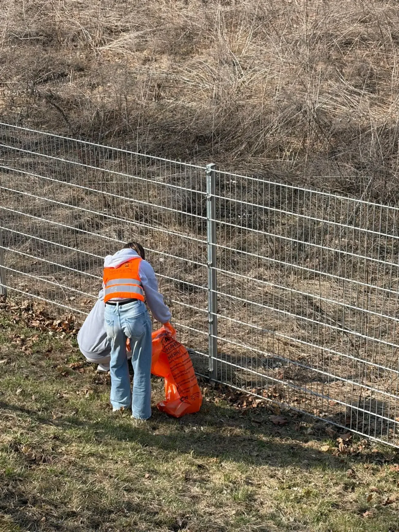 Eine Person mit einer Warnweste sammelt Müll an einem Zaun. Eine andere Person hockt hinter ihnen.