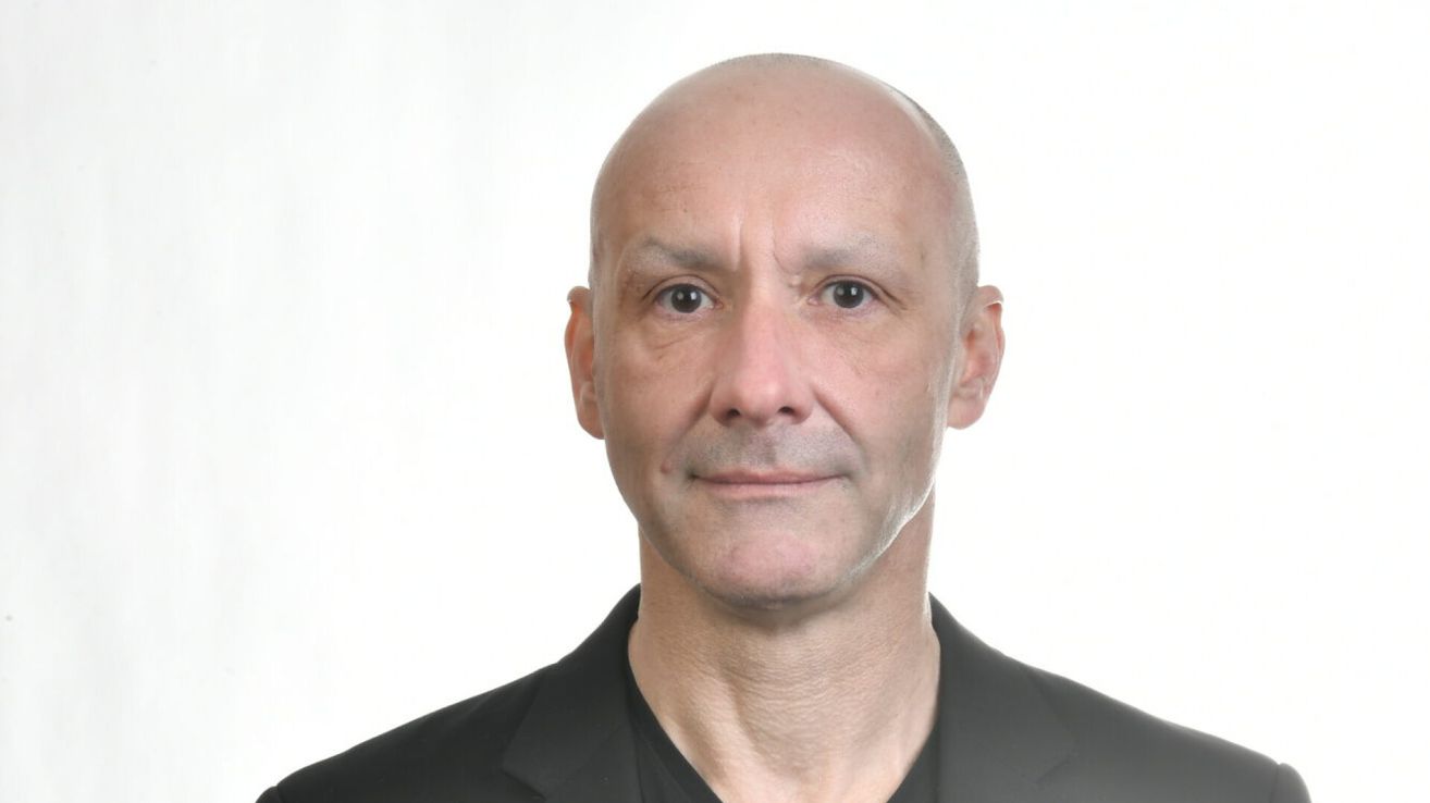 Ein mann mit kahlem kopf lächelt und posiert für ein foto. Er hat ein mal auf seiner stirn und trägt ein schwarzes hemd.