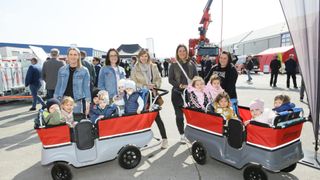 Eine Gruppe von Erwachsenen und Kindern an einem öffentlichen Ort. Mehrere Frauen schieben kleine Kinder in Spielzeugwagen. Im Hintergrund befinden sich Zelte und Rettungsfahrzeuge.