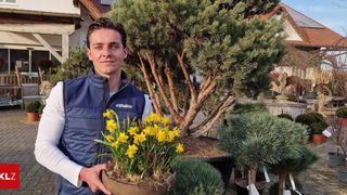 Ein Mann in einer blauen Weste hält einen Topf mit gelben Blumen. Er steht draußen, mit mehreren Bäumen im Hintergrund.