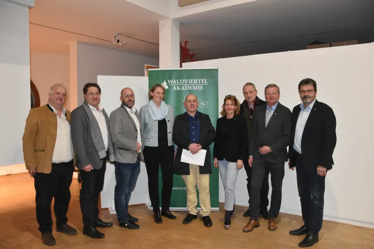Eine Gruppe von Personen in formeller Kleidung posiert für ein Foto vor einem grünen Banner mit der Aufschrift 'Waldviertel Akademie.' Der Mann in der Mitte hält ein Blatt Papier in der Hand. Die Umgebung scheint eine Indoor-Veranstaltung zu sein.