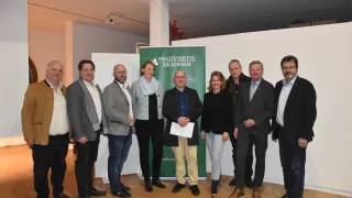 Eine Gruppe von Personen in formeller Kleidung posiert für ein Foto vor einem grünen Banner mit der Aufschrift 'Waldviertel Akademie.' Der Mann in der Mitte hält ein Blatt Papier in der Hand. Die Umgebung scheint eine Indoor-Veranstaltung zu sein.