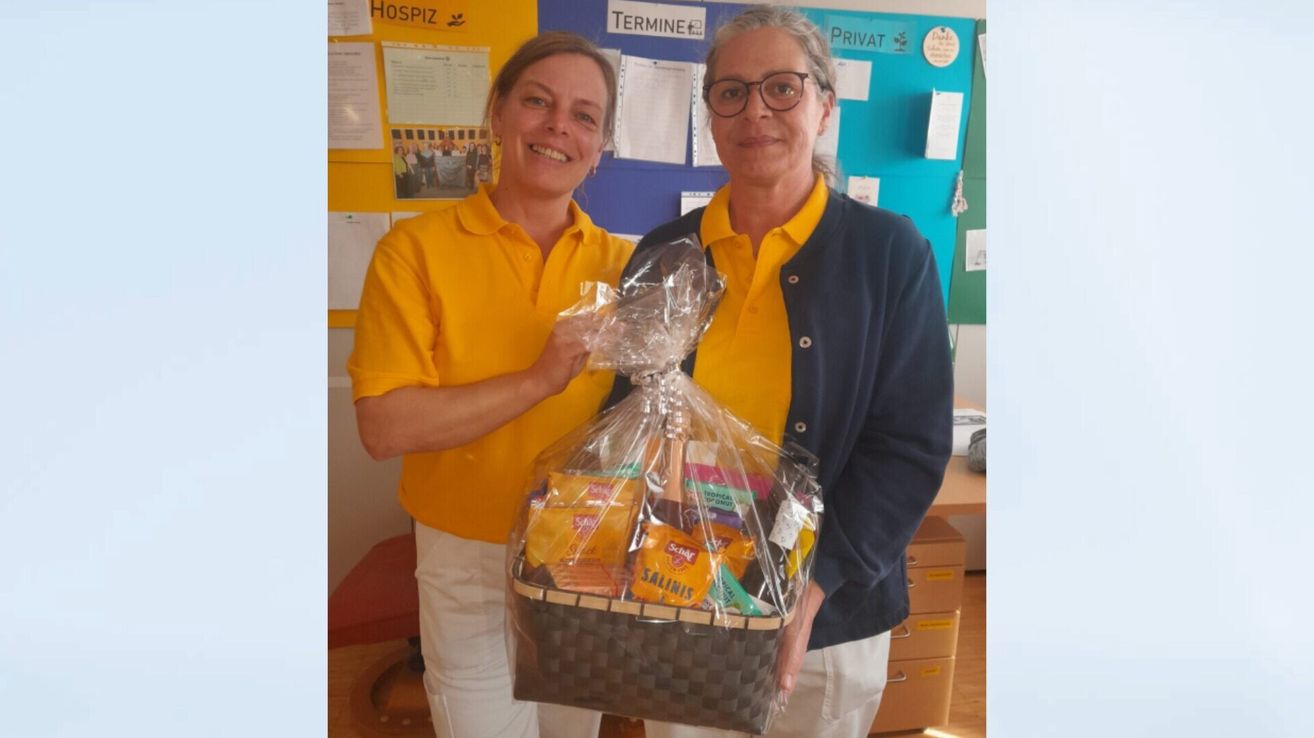 Zwei Frauen, beide in gelben Shirts, stehen nebeneinander und halten einen Geschenkkorb mit verschiedenen Snacks. Die Frau auf der linken Seite lächelt in die Kamera, während die andere zuschaut. Hinter ihnen ist ein Anschlagbrett mit Papieren und einem Plakat zu sehen.