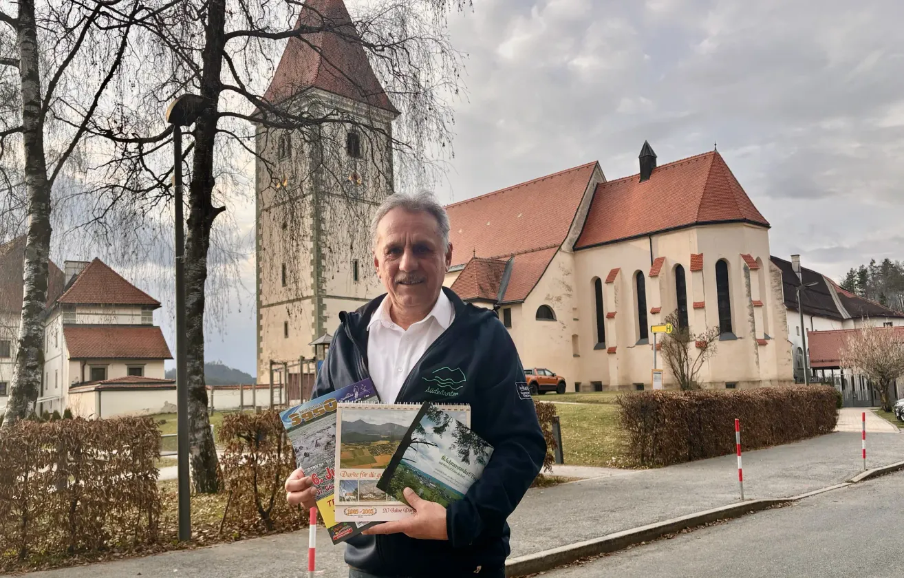 Ein Mann steht vor einer Kirche mit einem Turm und hält Broschüren in der Hand. Das Gebäude hat ein rotes Dach, und in der Nähe ist ein Auto geparkt.