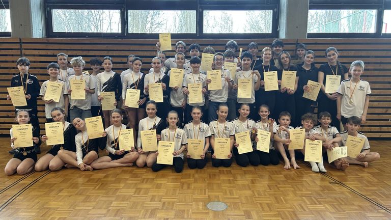 Eine Gruppe junger Athleten posiert für ein Foto in einer Turnhalle, hält Urkunden und Medaillen. Einige stehen, andere knien. Sie tragen weiße und schwarze Uniformen und Medaillen um den Hals.