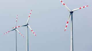 Ein Cluster von Windturbinen am blauen Himmel, jede Turbine hat drei Blätter und rote und weiße Streifen. Sie sind groß und metallisch.