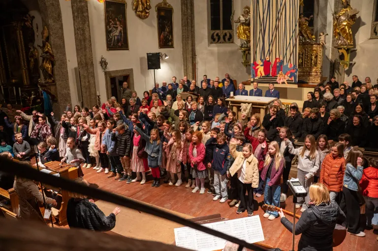 Ein Chor von Kindern in verschiedenen Outfits singt auf einer Bühne in einer Kirche. Ein Dirigent leitet von vorne, während ein Klavier auf der Bühne steht.