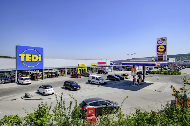 Ein Supermarktparkplatz mit mehreren geparkten Autos. Eine Tankstelle befindet sich rechts.