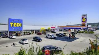 Ein Supermarktparkplatz mit mehreren geparkten Autos. Eine Tankstelle befindet sich rechts.