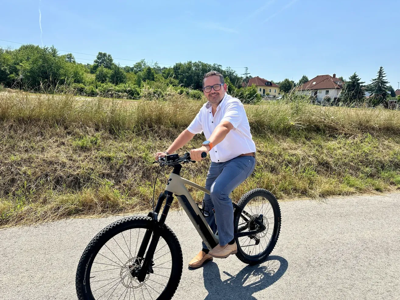 Ein Mann in einem weißen Hemd und einer Brille fährt auf einem schwarzen Fahrrad auf einer Straße mit Gras und Häusern im Hintergrund.