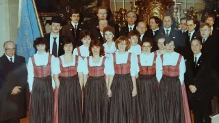 Eine Gruppe von Menschen in traditioneller Kleidung, die in einer Reihe stehen, einige mit Brille, möglicherweise für ein Foto posieren. Dahinter ein Weihnachtsbaum und aufwendige Dekorationen.