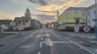 Eine ruhige Straße mit einem Sonnenuntergang in der Ferne, mit einigen Autos und einem Fußgänger im Vordergrund. Es gibt Straßenschilder und ein gelbes Gebäude auf der rechten Seite.