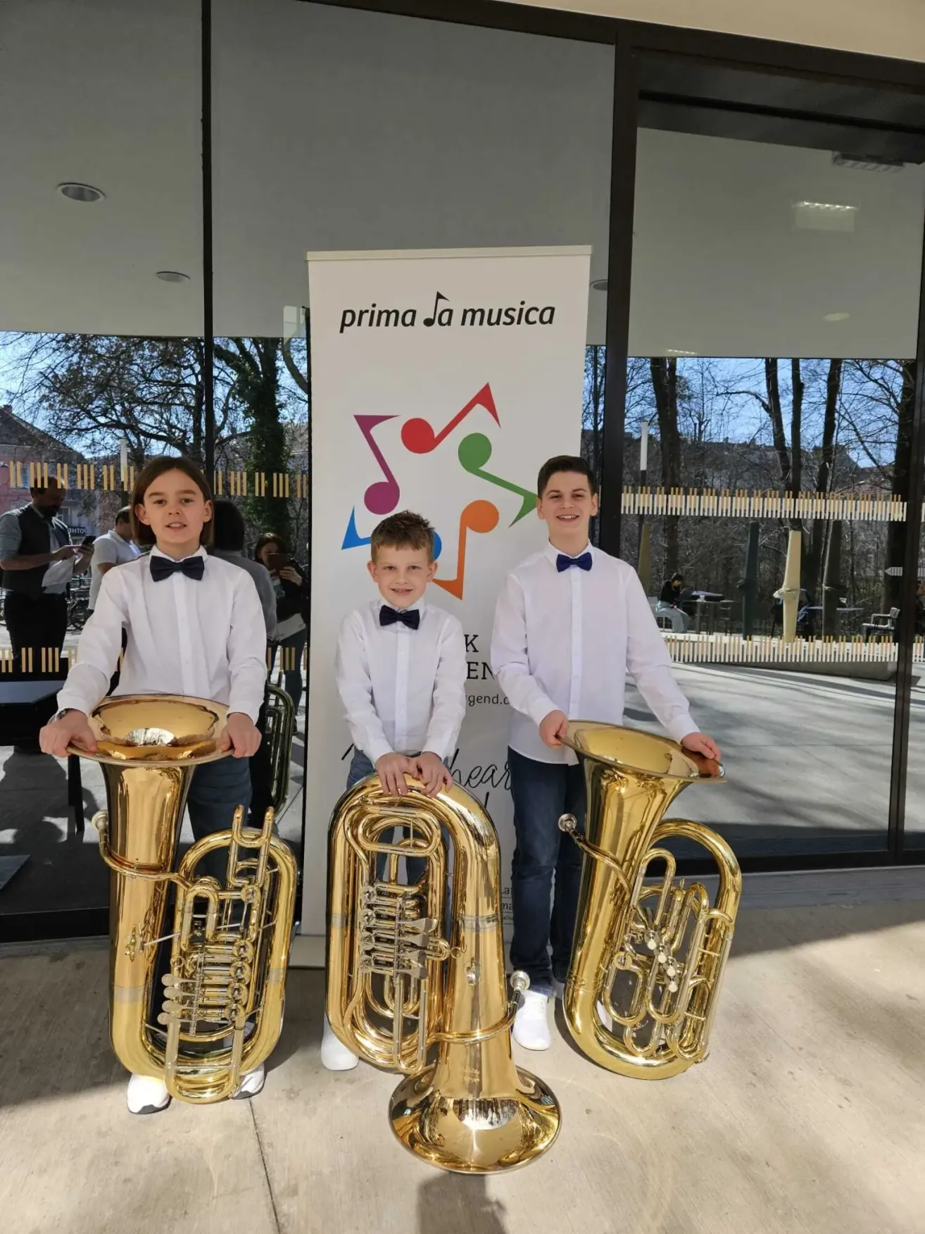 Drei Jungen in weißen Hemden und Fliegen posieren mit großen Blechblasinstrumenten vor einem Banner. Dahinter hält ein Mann mit Maske eine Kamera. Bäume und ein Gebäude sind im Hintergrund sichtbar.