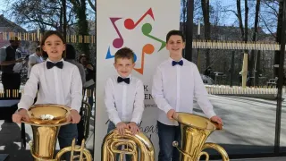 Drei Jungen in weißen Hemden und Fliegen posieren mit großen Blechblasinstrumenten vor einem Banner. Dahinter hält ein Mann mit Maske eine Kamera. Bäume und ein Gebäude sind im Hintergrund sichtbar.
