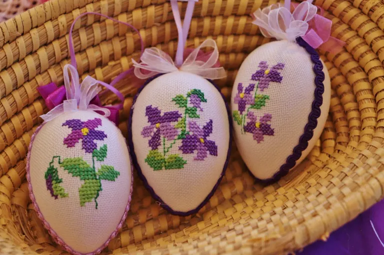 Drei kunstvoll bestickte weiße Eier mit lila Blumen und grünen Blättern, mit lila Bändern gebunden, liegen auf einem geflochtenen Korb.