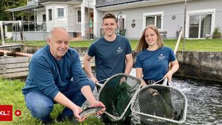 Drei Personen posieren am Bach mit Angelkörben, mit einem Haus und Zaun im Hintergrund.