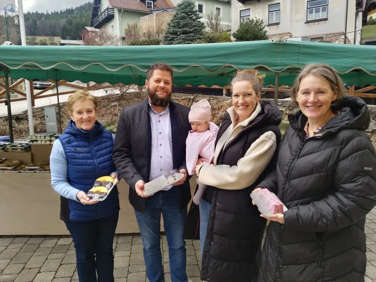 Vier Menschen stehen in einer Reihe, lächeln und halten verschiedene Gegenstände. Ein Mann hält ein Baby, und sie stehen vor einem grünen Zelt.