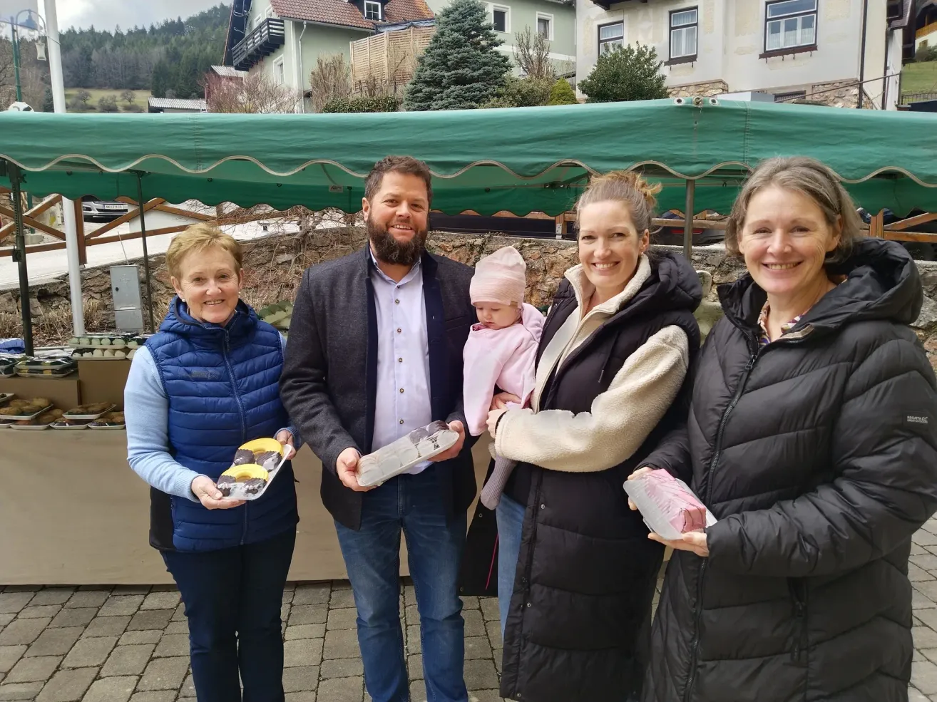 Vier Menschen stehen in einer Reihe, lächeln und halten verschiedene Gegenstände. Ein Mann hält ein Baby, und sie stehen vor einem grünen Zelt.