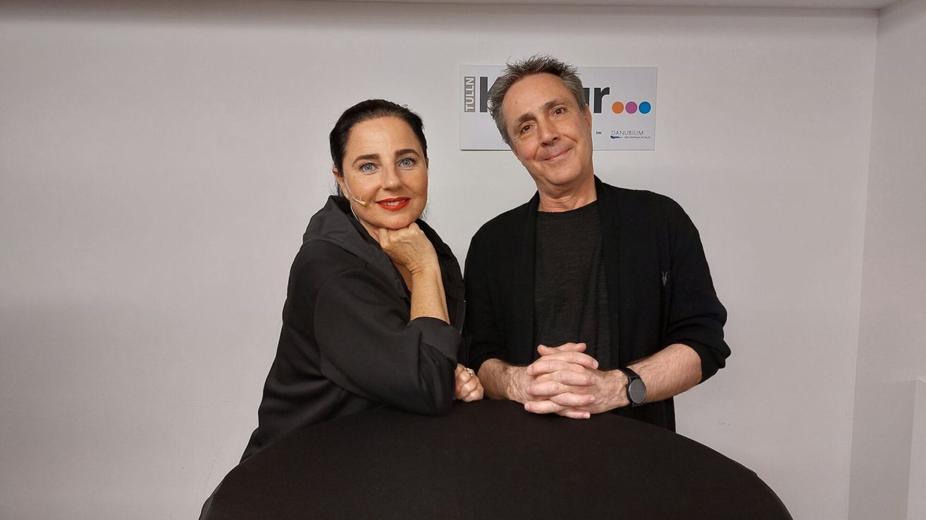 Ein Mann und eine Frau sitzen lächelnd an einem runden Tisch. Die Frau trägt ein Headset. Dahinter steht ein Schild mit den Aufschriften 'Kr Tulln' und 'Danubium'.