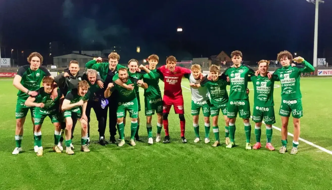 Fußballspieler, alle mit grünen Trikots und verschiedenen Sponsorenlogos, feiern einen Sieg auf dem Feld. Ein Mann in Rot steht in der Mitte.