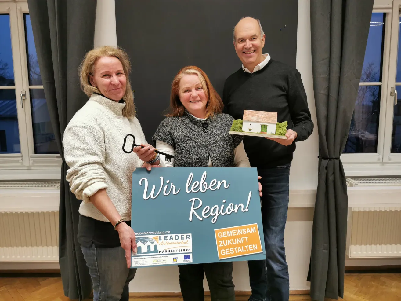 Drei Menschen stehen eng beieinander und halten einen Schlüssel und ein Modellhaus. Das Schild sagt 'Wir leben Region!' Sie scheinen für ein Foto zu posieren.