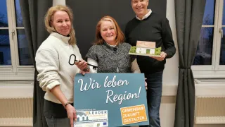Drei Menschen stehen eng beieinander und halten einen Schlüssel und ein Modellhaus. Das Schild sagt 'Wir leben Region!' Sie scheinen für ein Foto zu posieren.