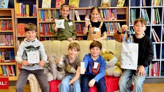 Eine Gruppe von Kindern, die lächeln und Gegenstände halten, sitzen auf einer Couch in einer Bibliothek. Sie sind von Plüschtieren und Büchern umgeben.
